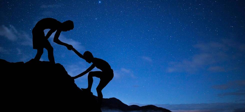 Ein Sternenhimmel bei Nacht im Hintergrund, im Vordergrund die Silhouetten von zwei Personen, die einen Berg hinaufsteigen.