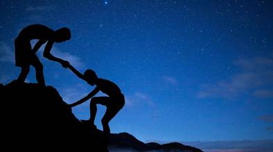 Ein Sternenhimmel bei Nacht im Hintergrund, im Vordergrund die Silhouetten von zwei Personen, die einen Berg hinaufsteigen.