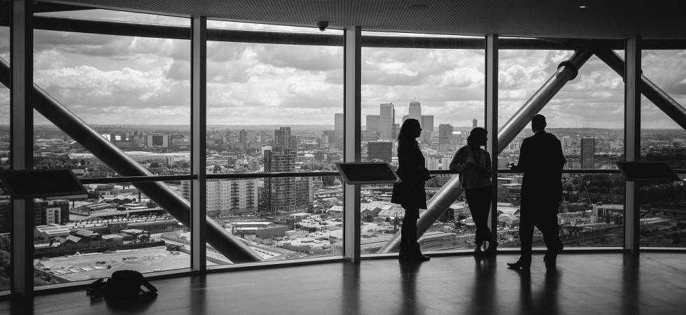 Schwarz-weiß-Bild von drei Menschen, die sich mit dem Rücken zu einer Glasfront unterhalten. Hinter ihnen sieht man die Skyline der Stadt. Links auf dem Boden liegt ein Rucksack